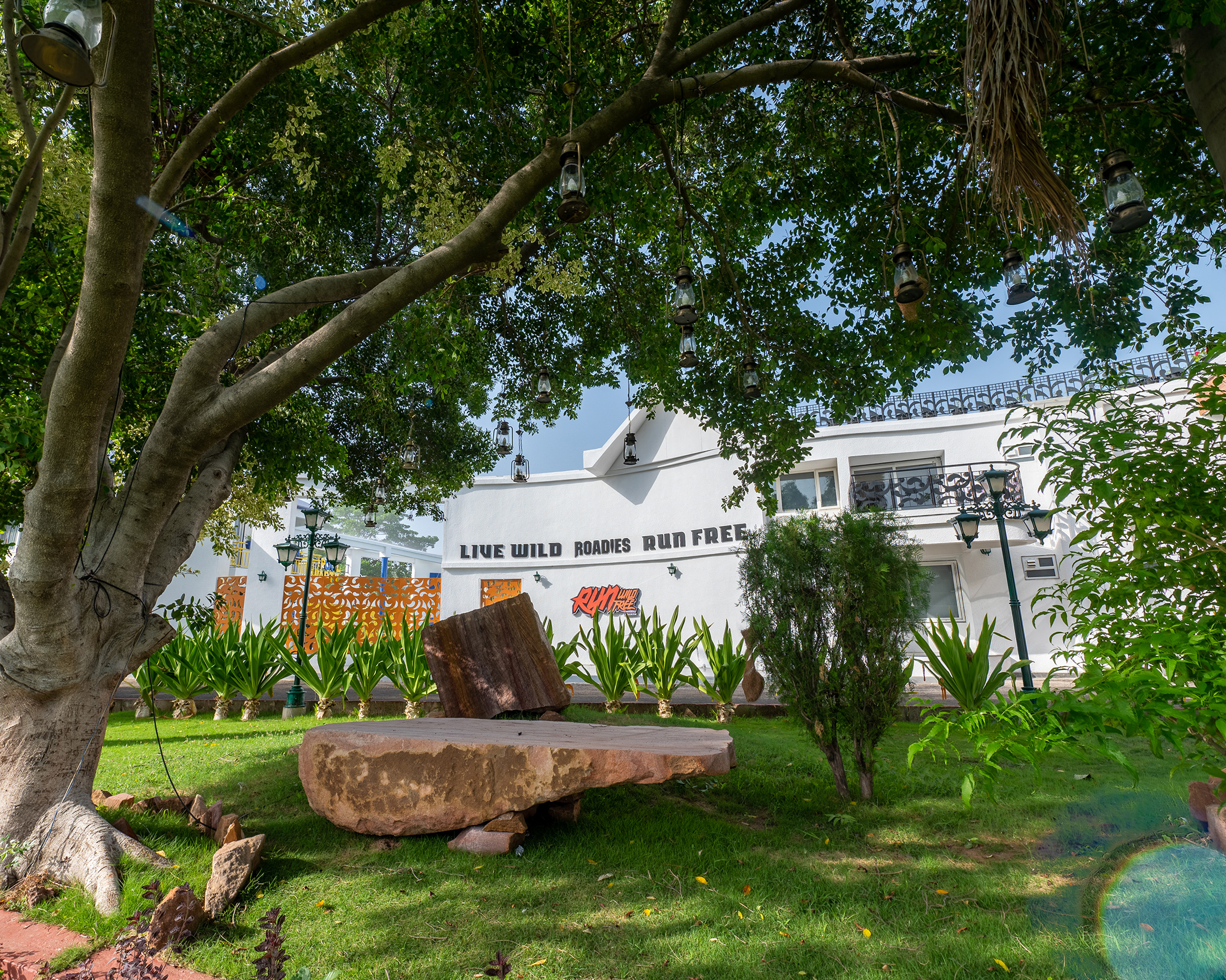 Roadies Rostel garden seating area under large tree with hanging lanterns and resort wall in background