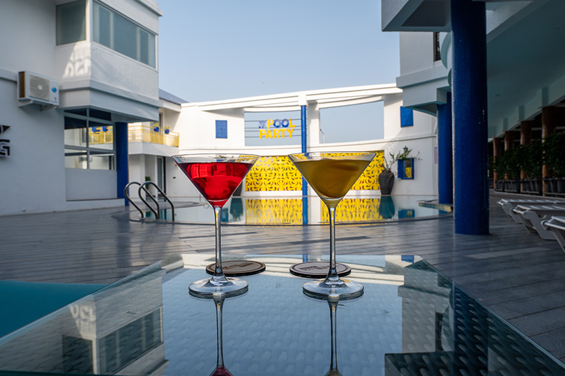 Two vibrant mocktails placed on a glass table beside the pool at Roadies Rostel with a pool party backdrop