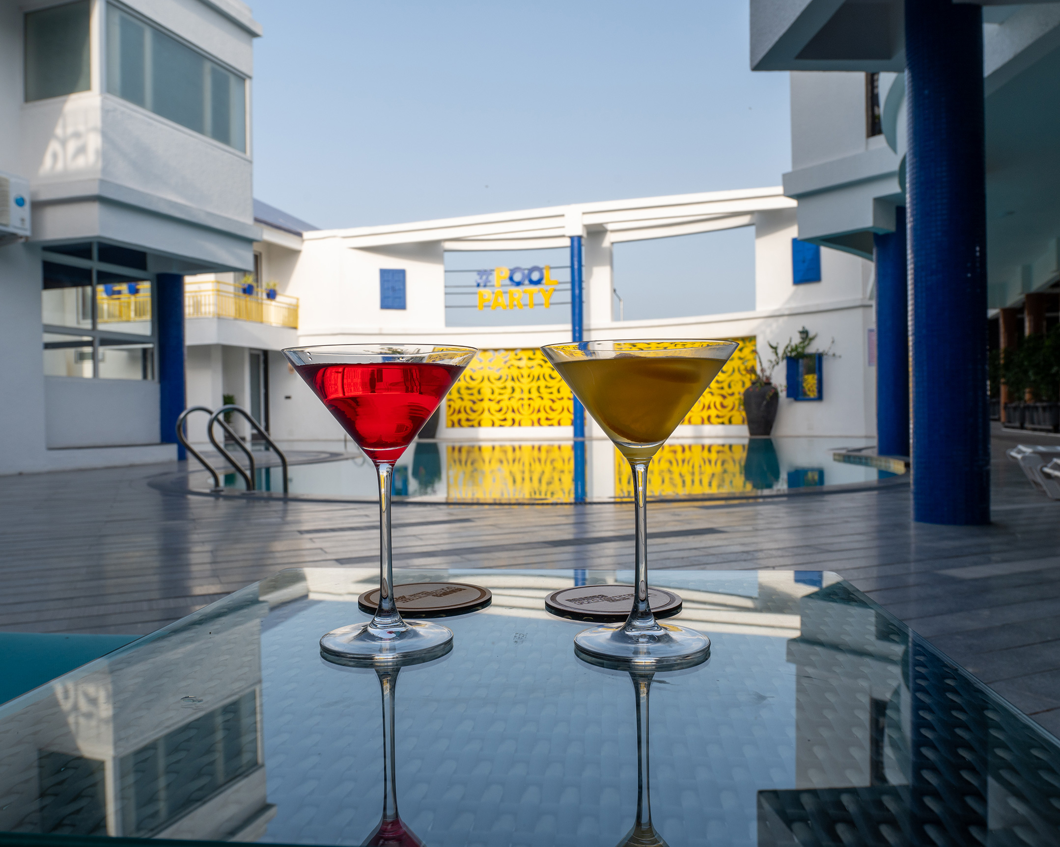 Two vibrant mocktails placed on a glass table beside the pool at Roadies Rostel with a pool party backdrop